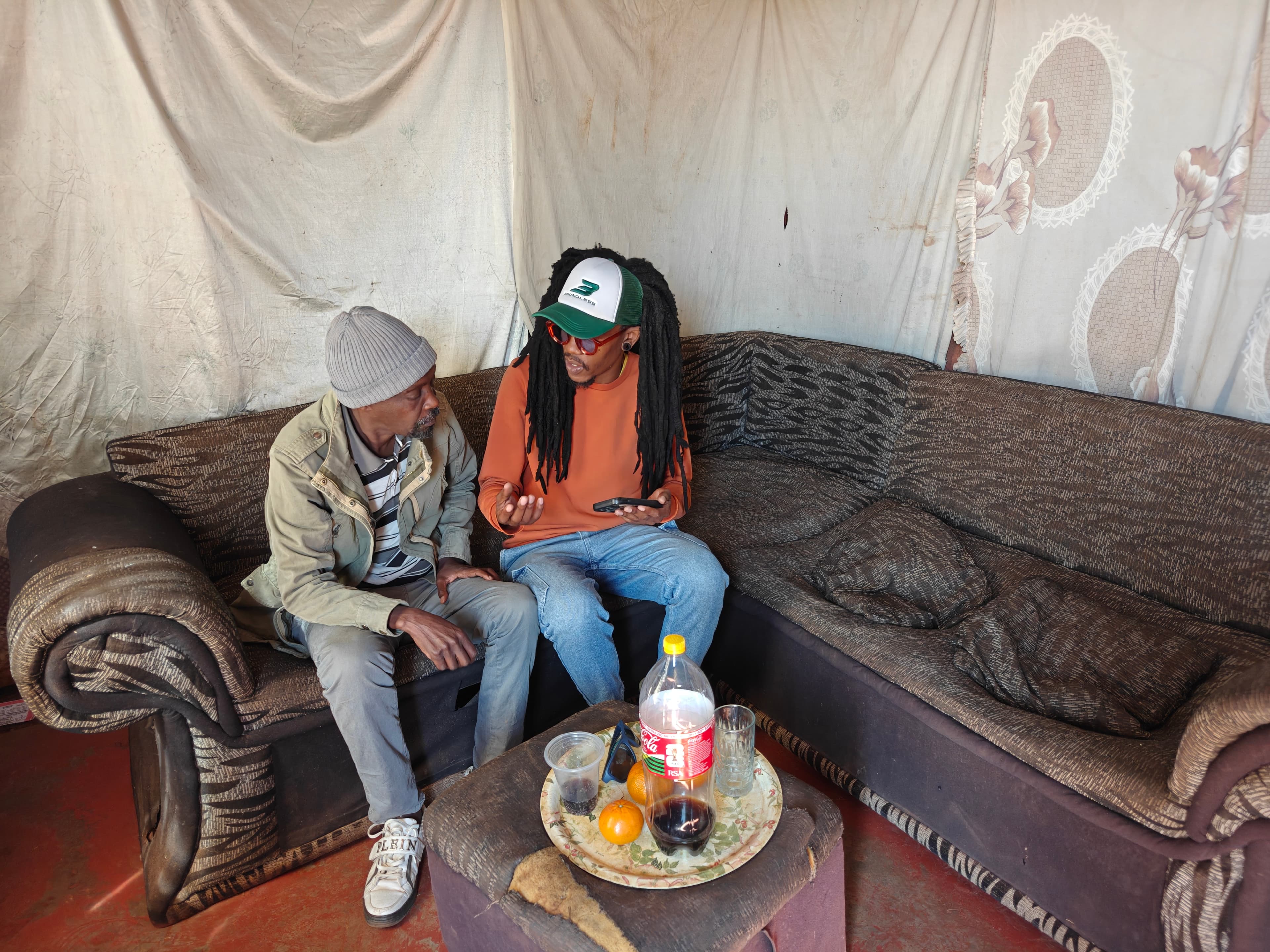 Two men sit on a patterned sofa in a room lined with fabric sheets, deep in conversation. One wears a grey beanie and olive jacket; the other, with long dreadlocks, a cap and an orange jumper, gestures while holding a phone. A tray with a bottle of cola, cups and oranges sits on a low table in front of them.